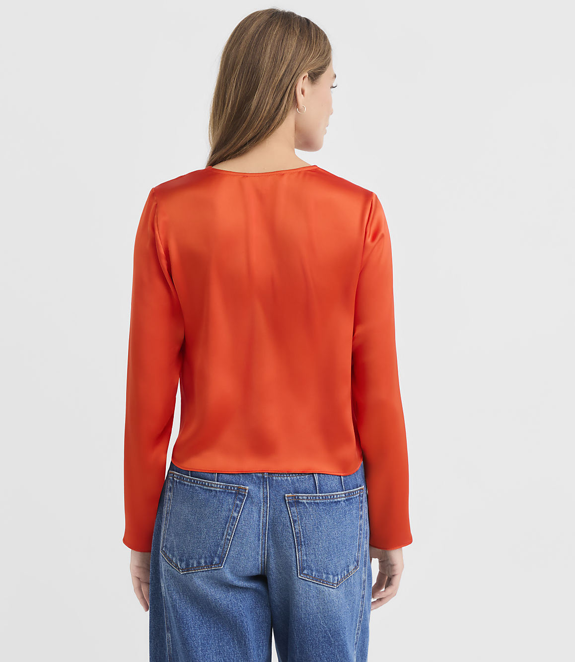 A woman in a vibrant orange satin top and blue jeans stands against a plain white background.