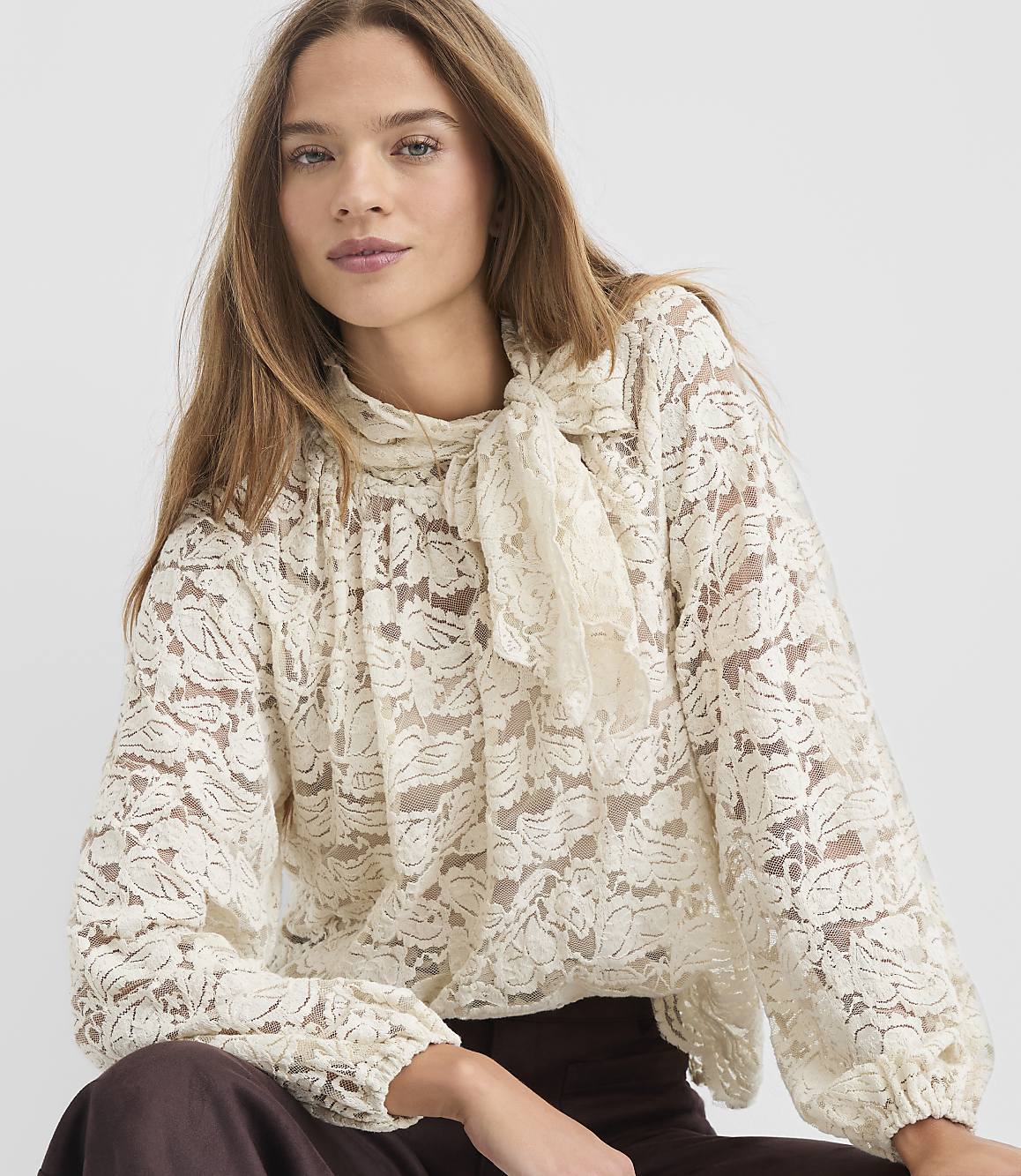 A woman with long brown hair wears a cream-colored lace blouse with a pussy-bow tie, against a plain white background.