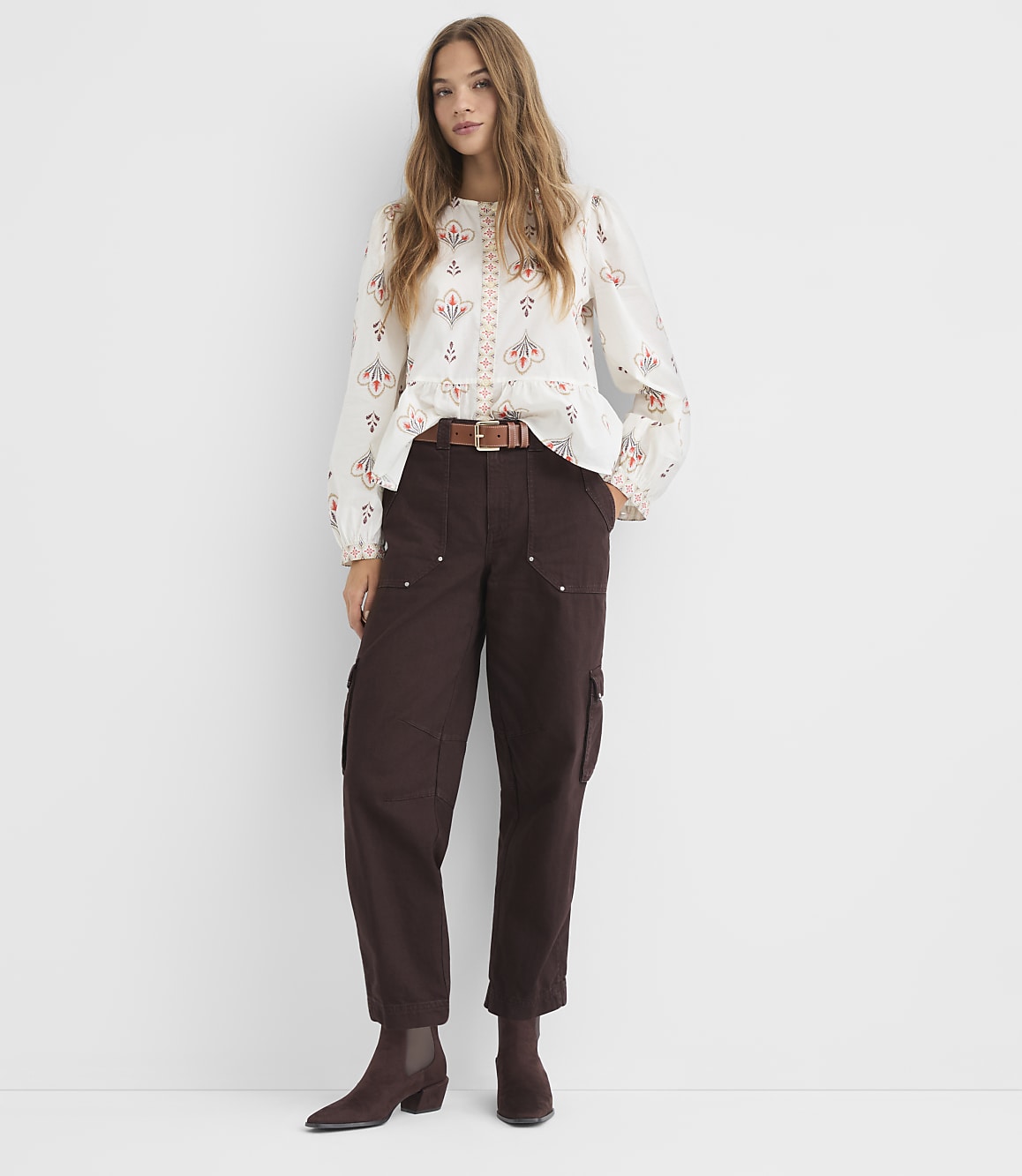 A woman in a patterned blouse and brown cargo pants stands against a plain white background.