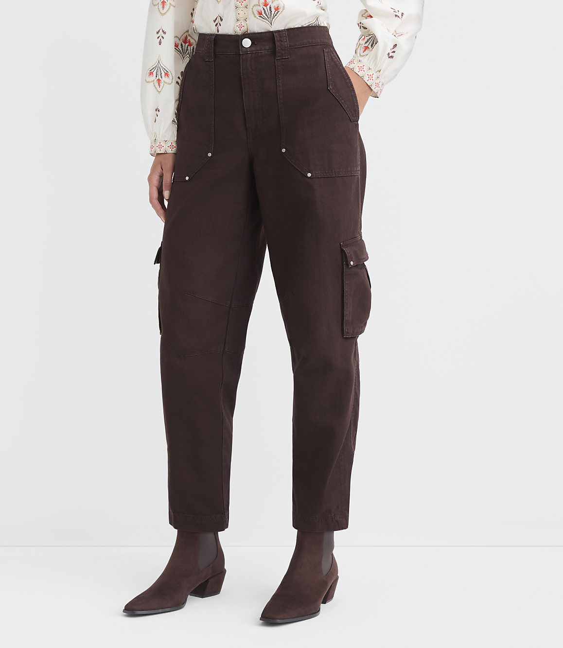 A person stands wearing brown cargo pants and a patterned blouse, with a plain white background.