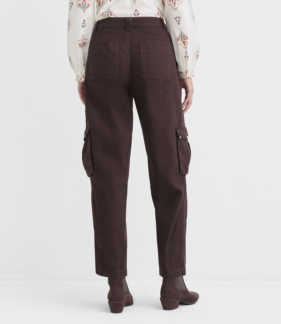 A person wearing brown cargo pants and a patterned blouse stands against a plain white background.
