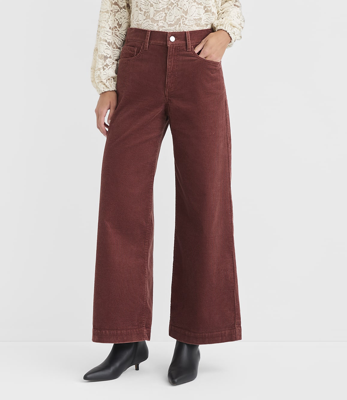 A person in rust-colored corduroy pants and a lace top stands against a plain white background.