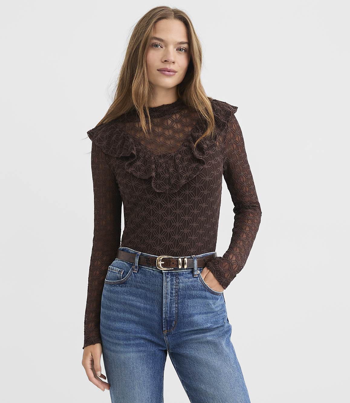 A woman in a brown ruffled top and blue jeans stands against a textured white background.