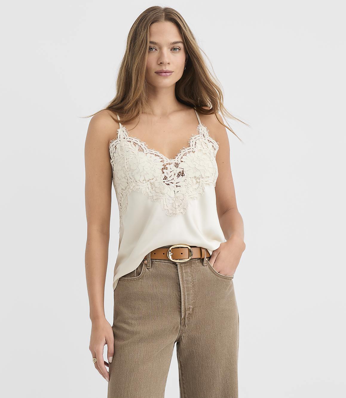 A woman in a cream lace camisole and brown pants stands against a plain white background.