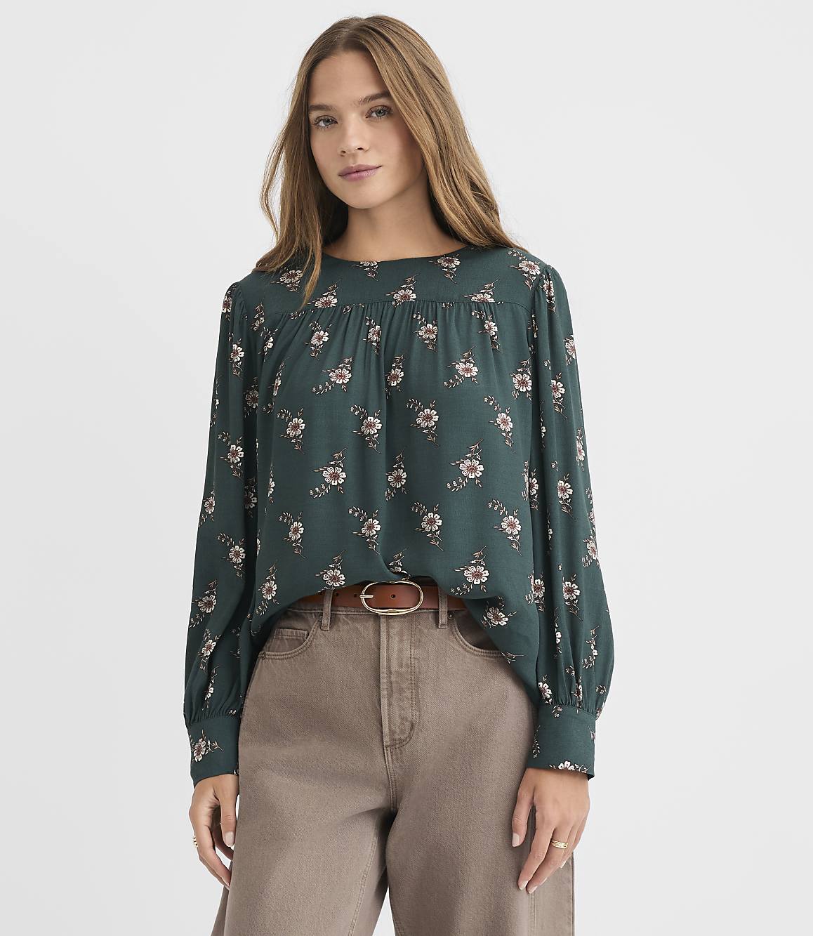 A woman in a dark green floral blouse and brown pants stands against a plain white background.