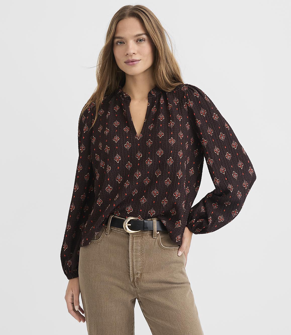 A woman in a patterned blouse and tan pants stands against a plain white background.