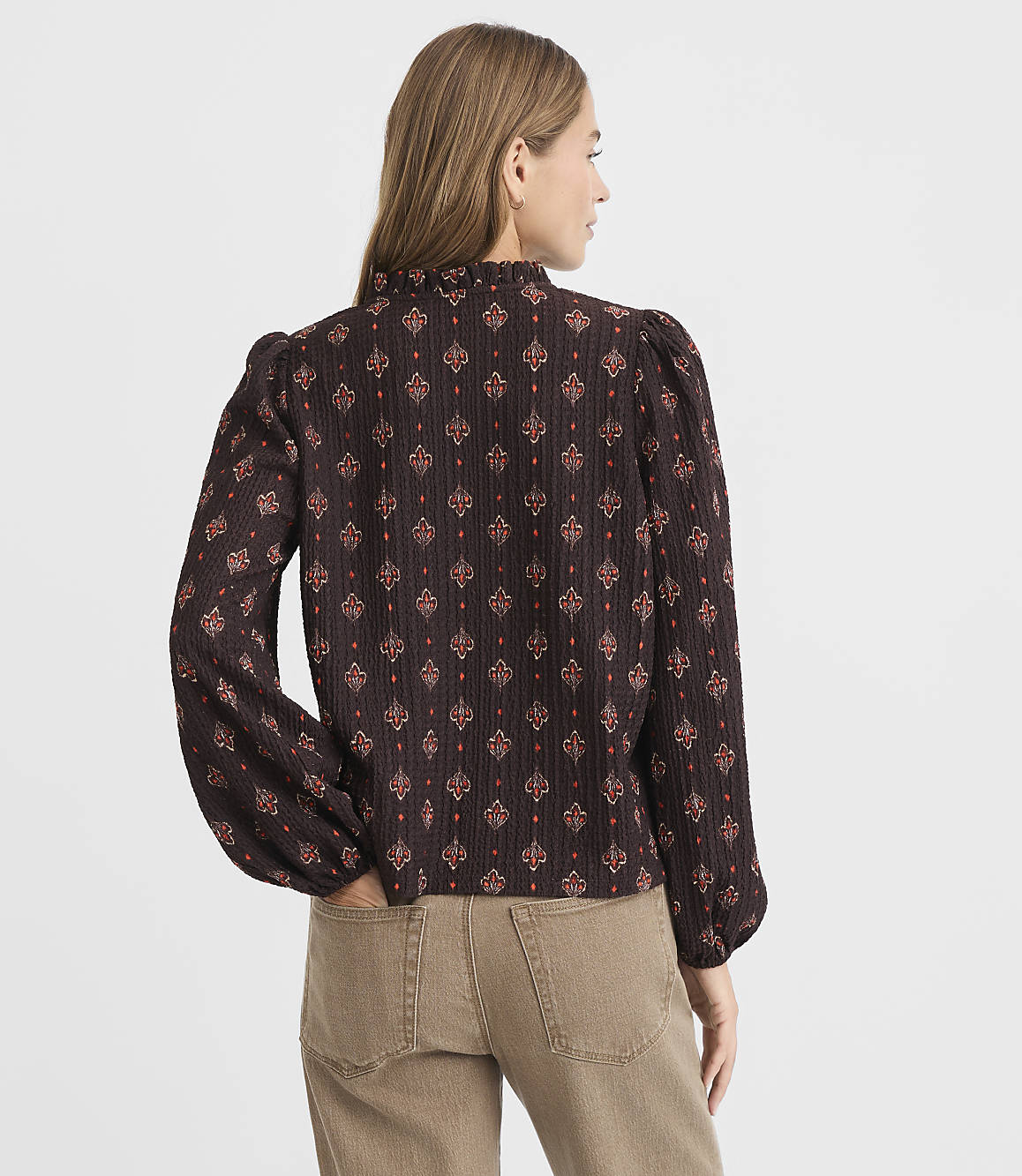A woman in a patterned blouse and tan pants stands against a plain white background.