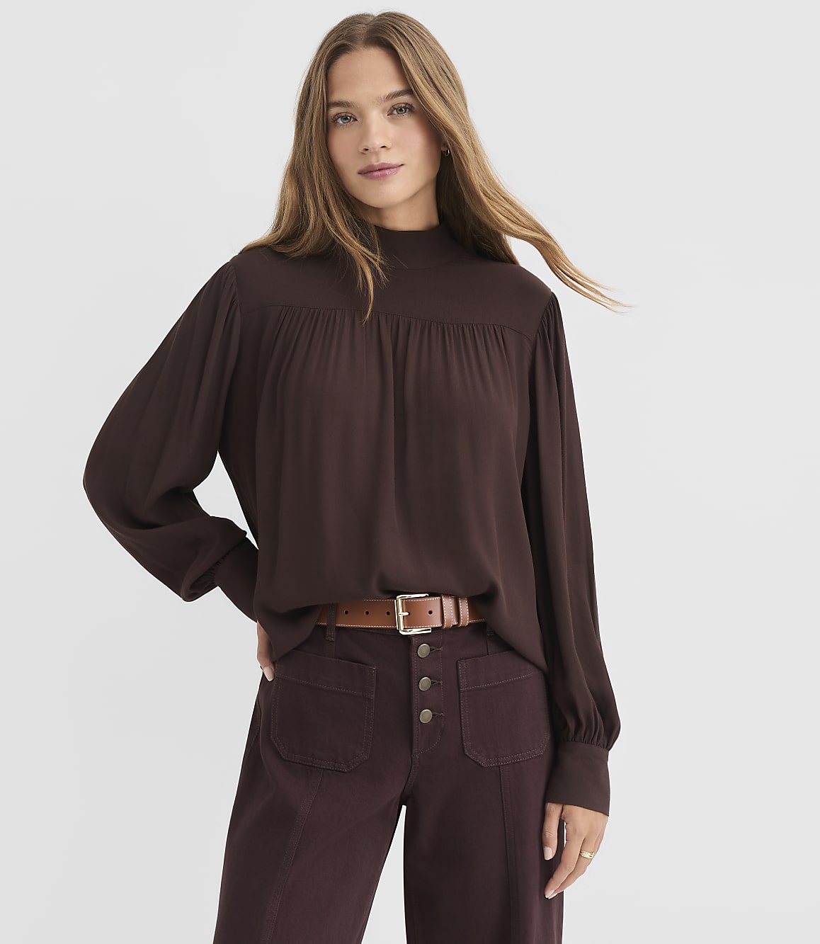 A woman in a brown mock-neck blouse and maroon pants stands against a plain white background.
