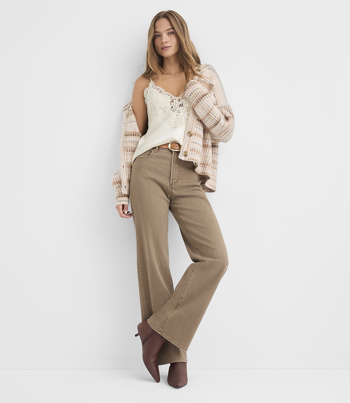 A young woman in a cream lace camisole, a plaid cardigan, and brown wide-leg jeans stands against a plain white background.