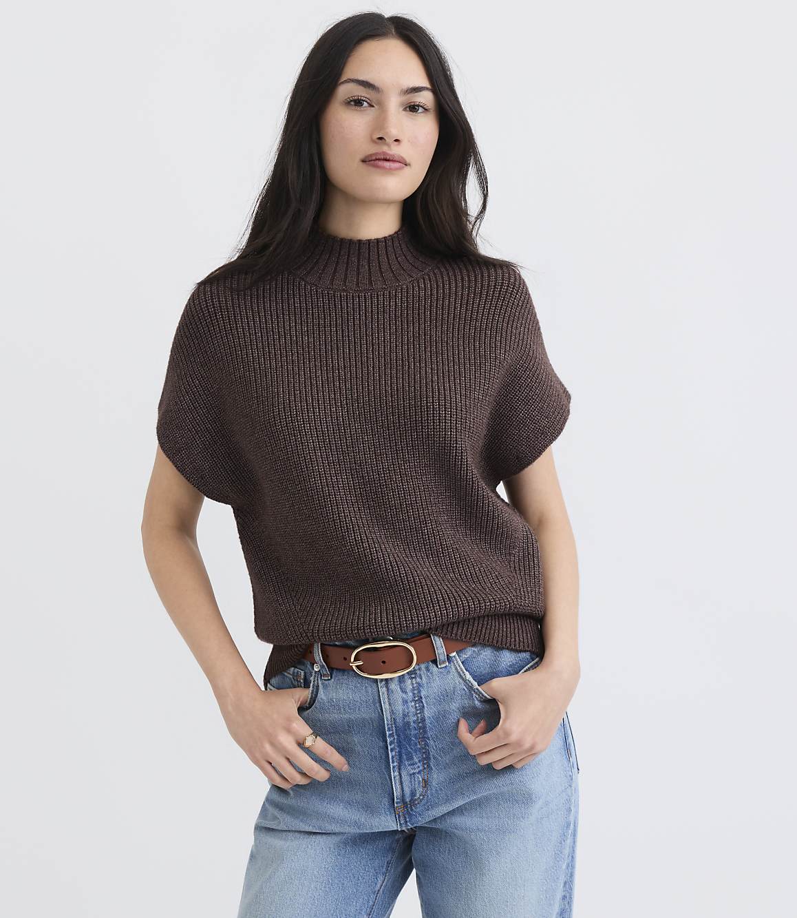 A woman in a brown mock-neck sweater and blue jeans stands against a plain white background.
