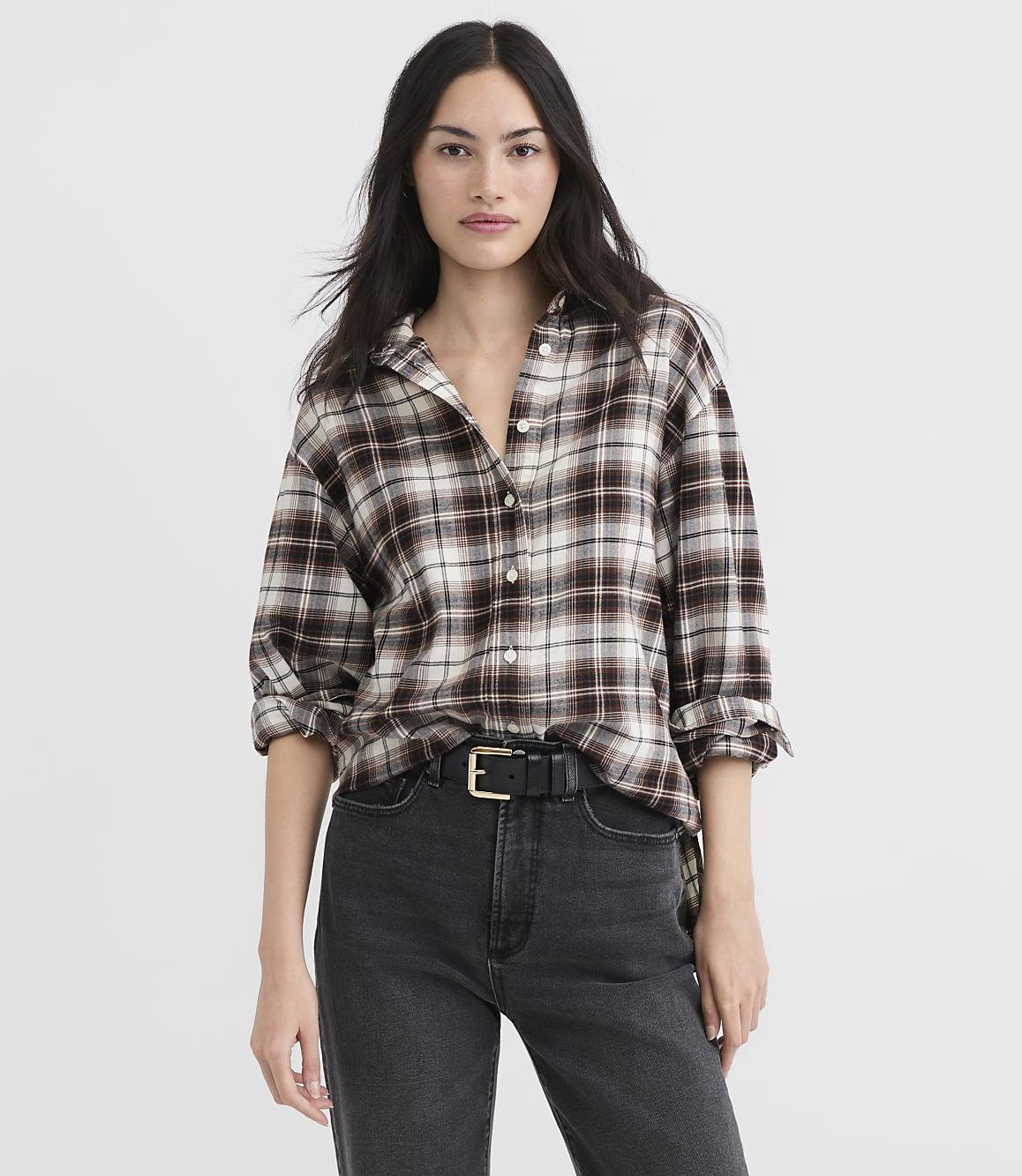 A woman in a brown plaid shirt and black jeans stands against a plain white background.