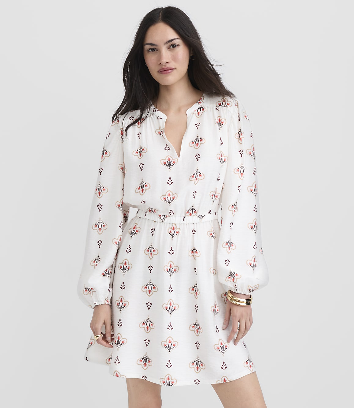A woman in a white patterned dress stands against a plain white background.
