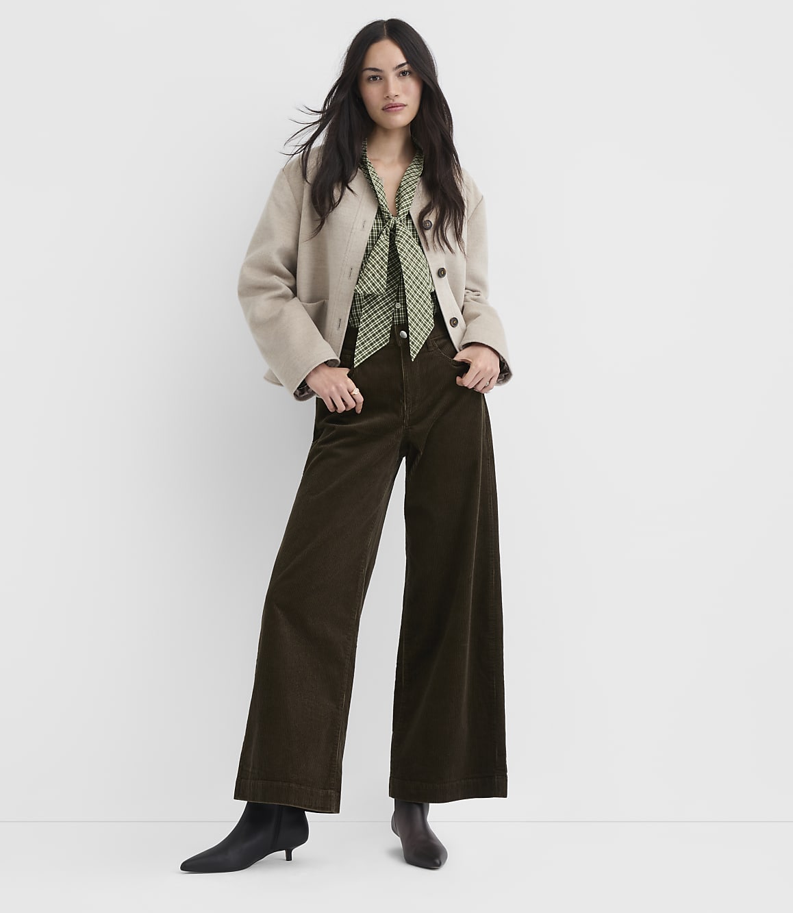 A woman in a beige jacket, green patterned blouse, and brown corduroy pants stands against a plain white background.