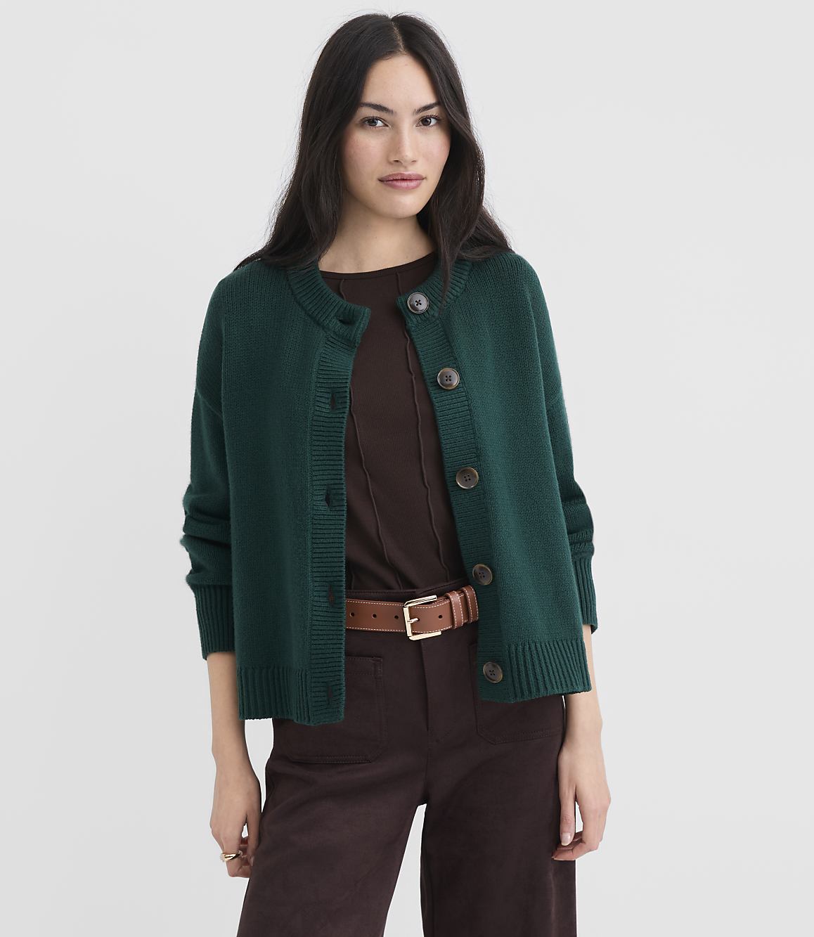 A woman in a green cardigan and brown pants stands against a plain white background.