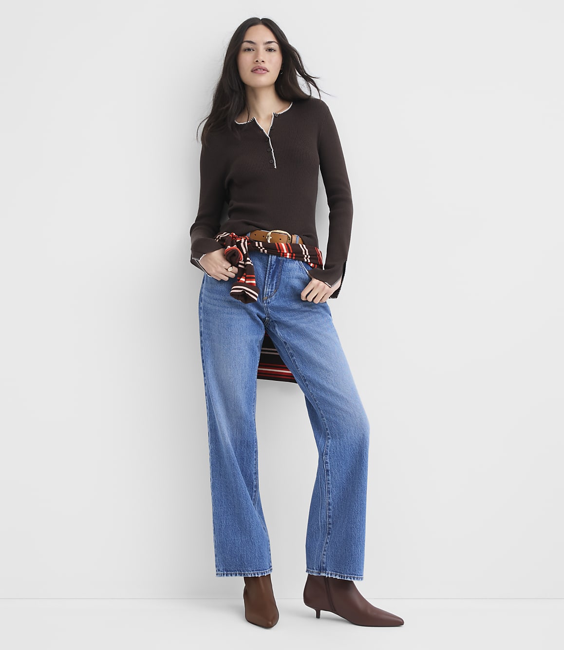 A woman in a brown henley and blue jeans stands against a plain white background, with a plaid shirt tied around her waist.