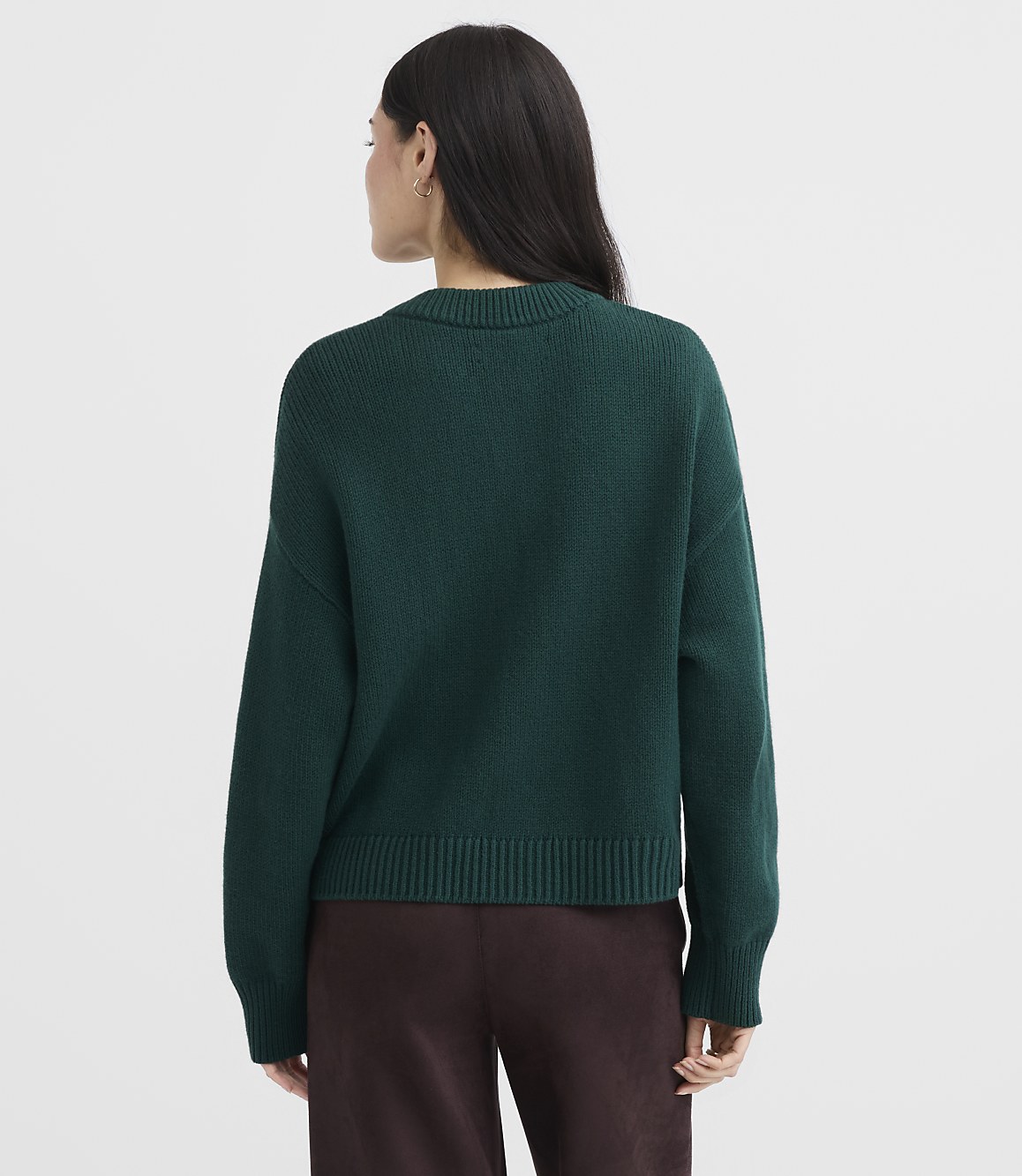 A woman in a dark green sweater and brown pants stands against a plain white background.