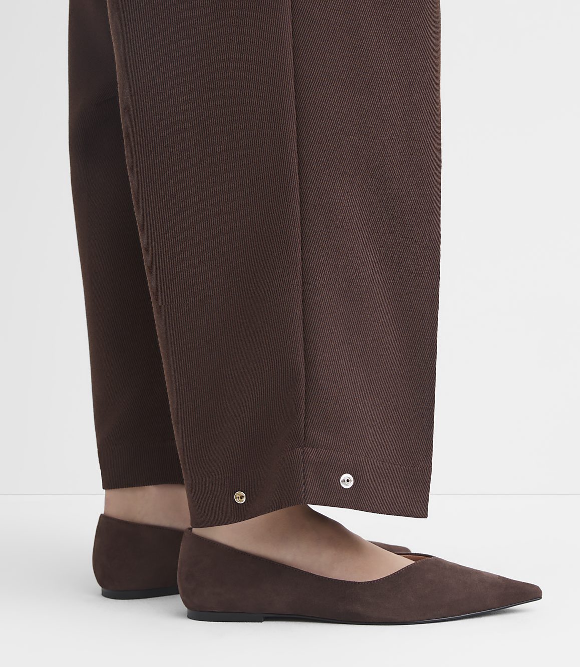 Brown suede flats peek out from beneath the hem of brown trousers adorned with silver buttons.