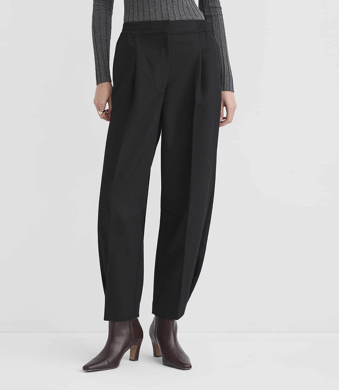 A person stands in black pleated trousers and brown ankle boots against a plain white background.