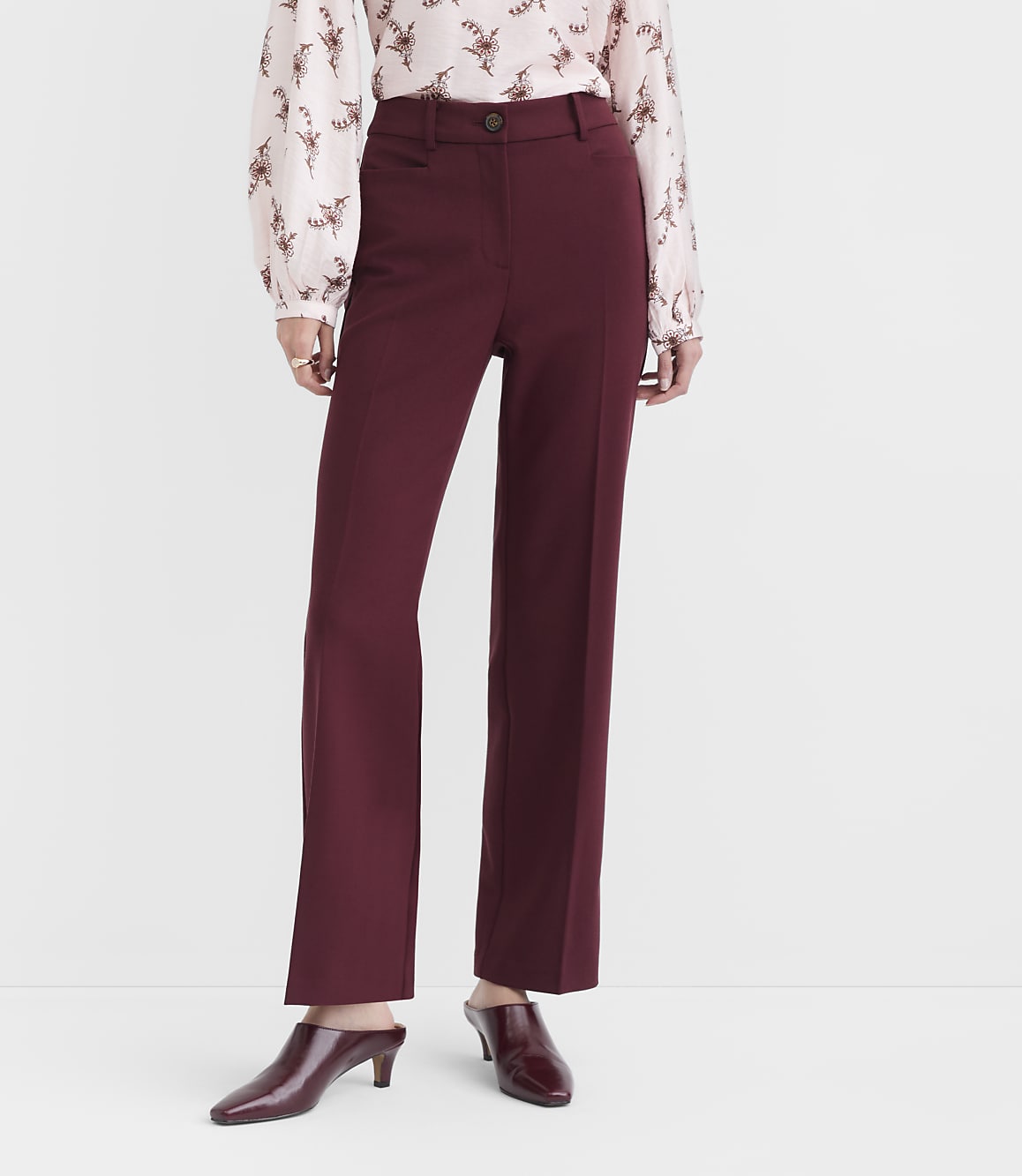 A person in a patterned blouse and burgundy trousers stands against a plain, light gray background.