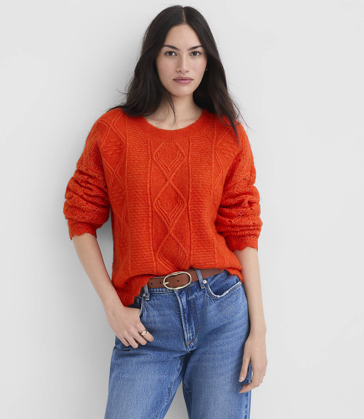 A woman in a bright orange cable-knit sweater and blue jeans stands against a plain white background.