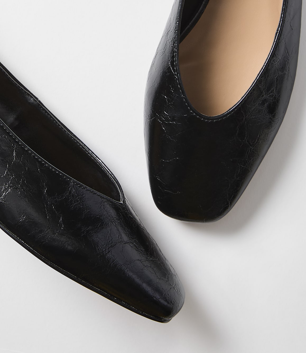 Two black, square-toed flats with a slightly crinkled texture are positioned on a plain white background.