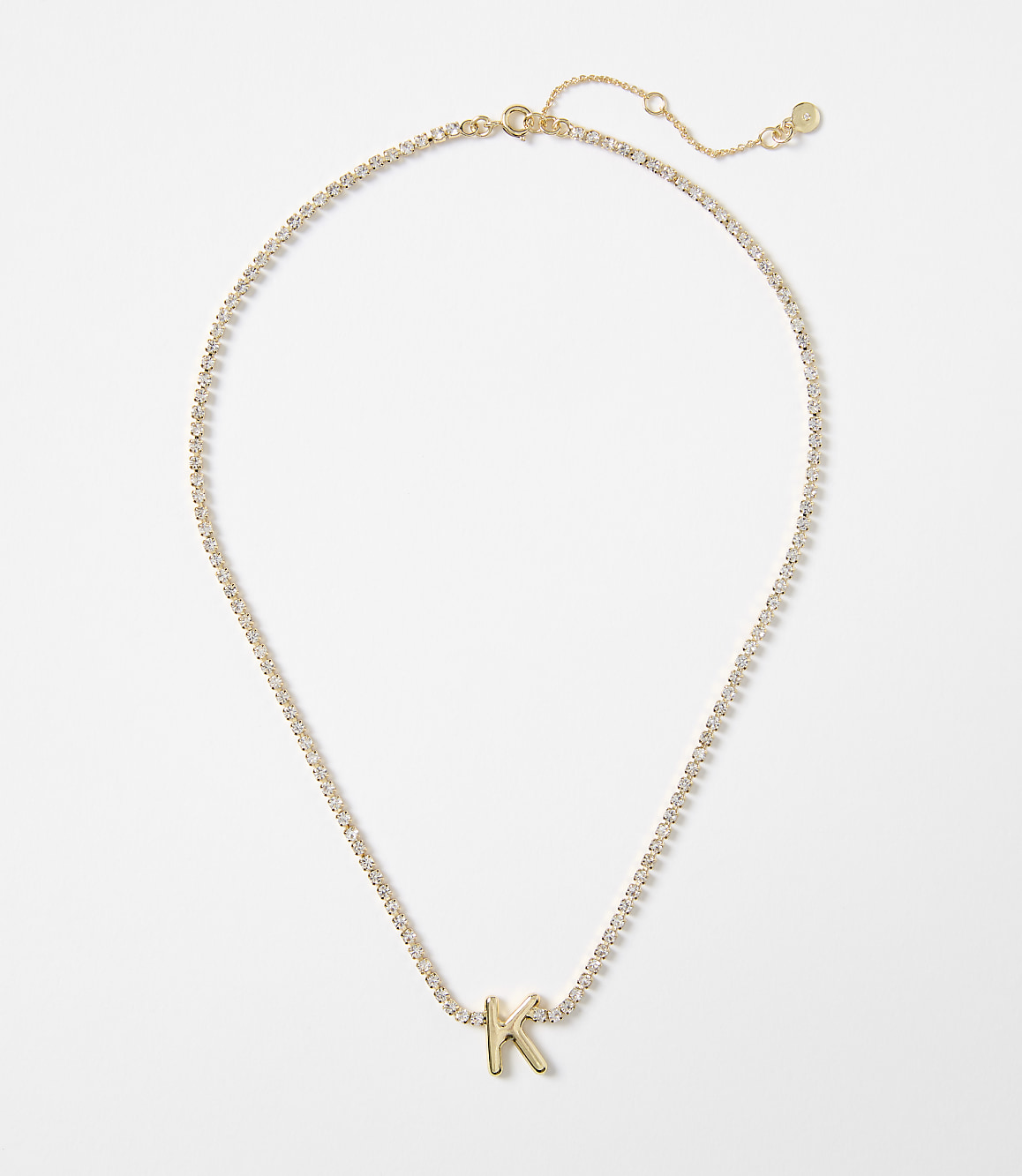 A delicate gold chain with a sparkling crystal tennis necklace design is adorned with a single gold letter "K" charm against a plain white background.
