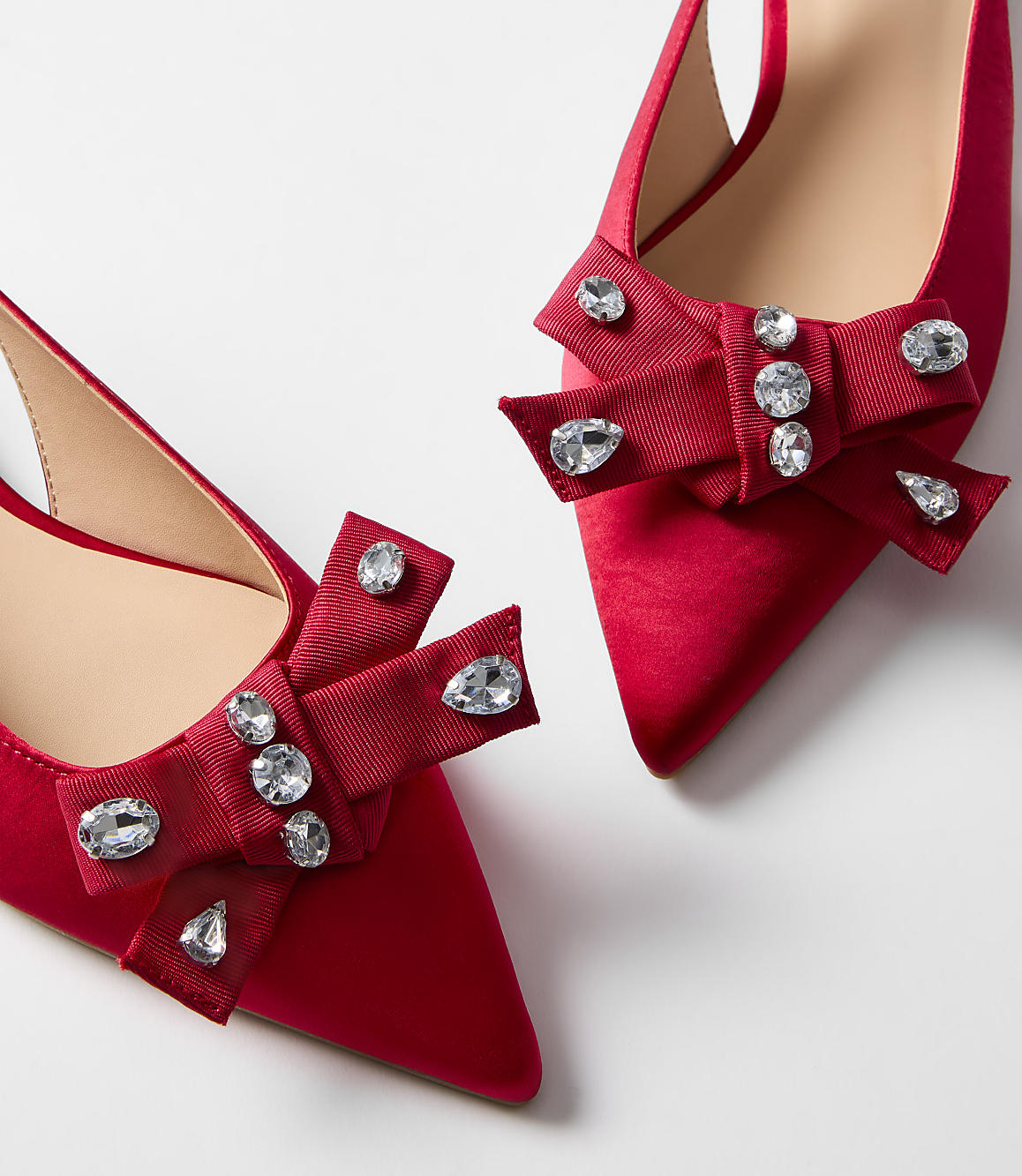 Two red satin pointed-toe heels with jeweled bows are displayed against a plain white background.