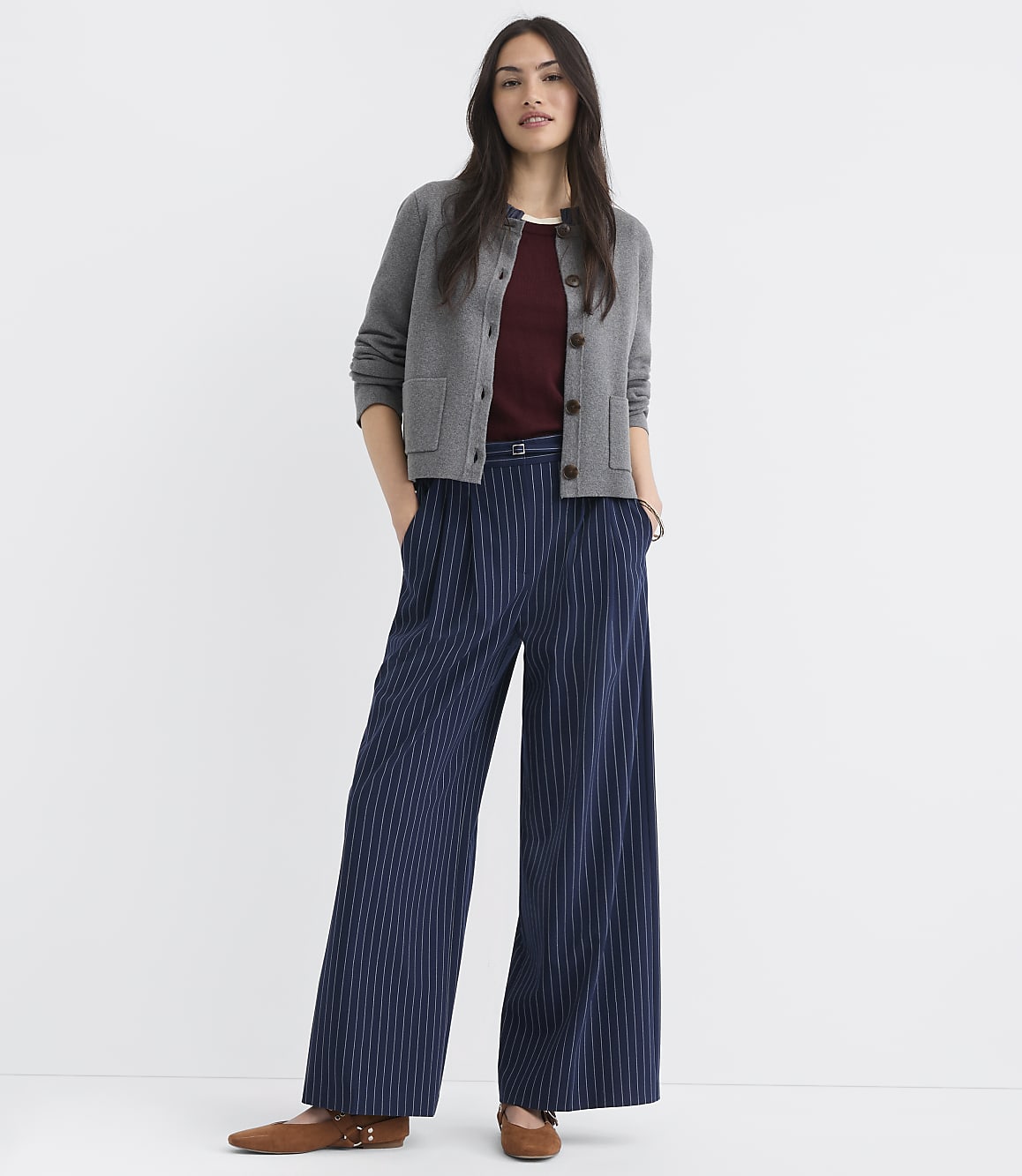 A woman in a gray cardigan and pinstripe pants stands against a plain white background.