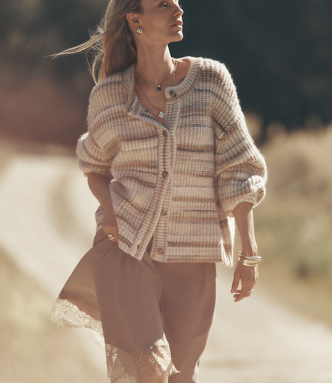 A woman in a striped cardigan and slip dress walks down a dirt road with trees in the background.