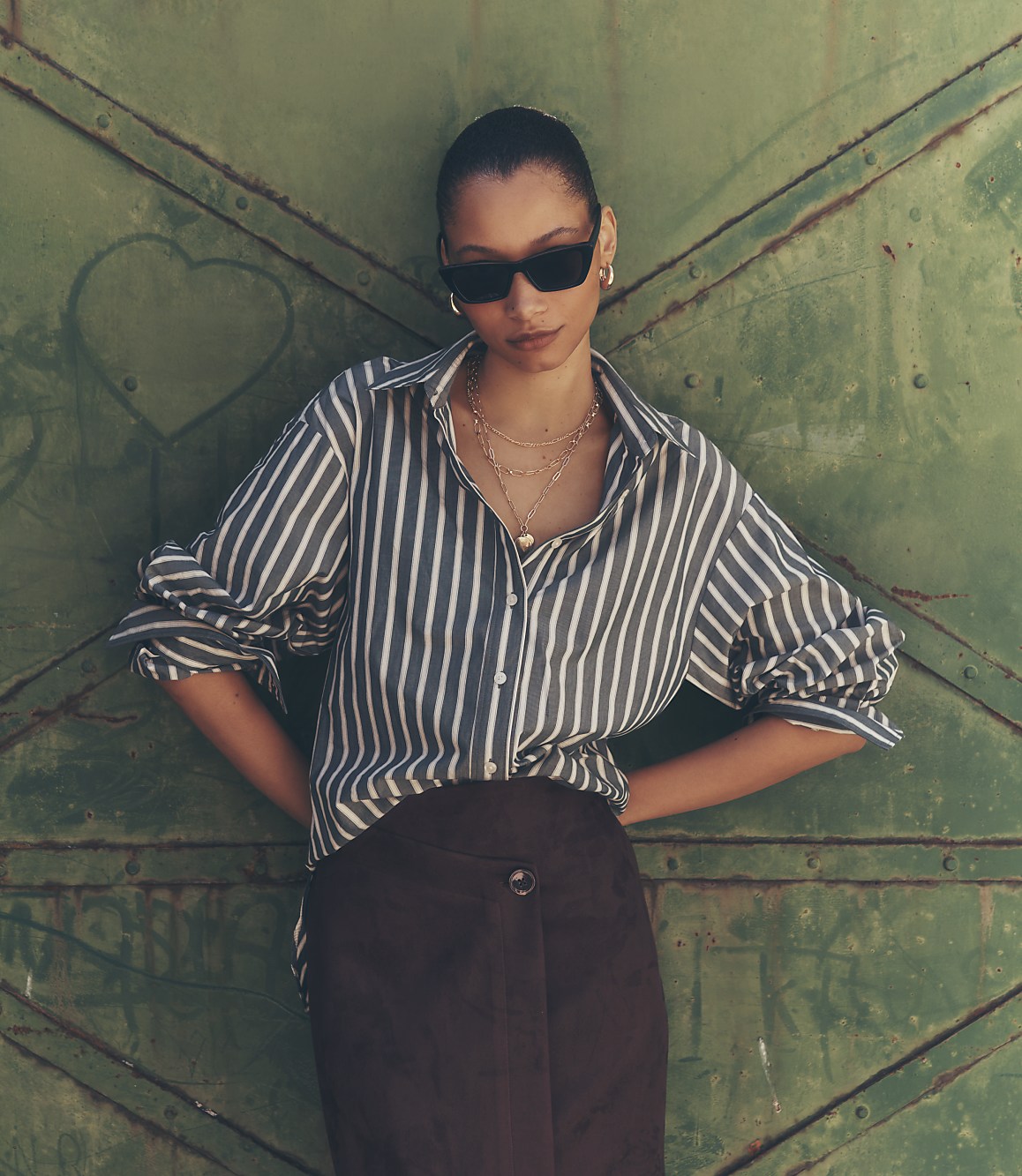 A woman in a striped shirt and dark skirt stands against a green, textured metal door.