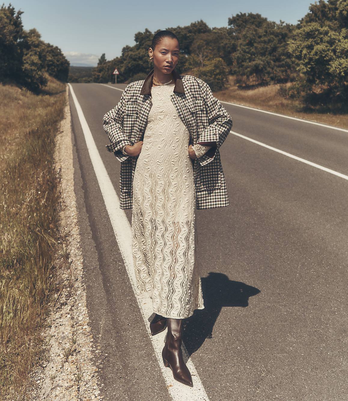 A woman in a patterned jacket and lace dress stands on an empty asphalt road with grassy verges, leading towards a tree-lined horizon under a clear sky.
