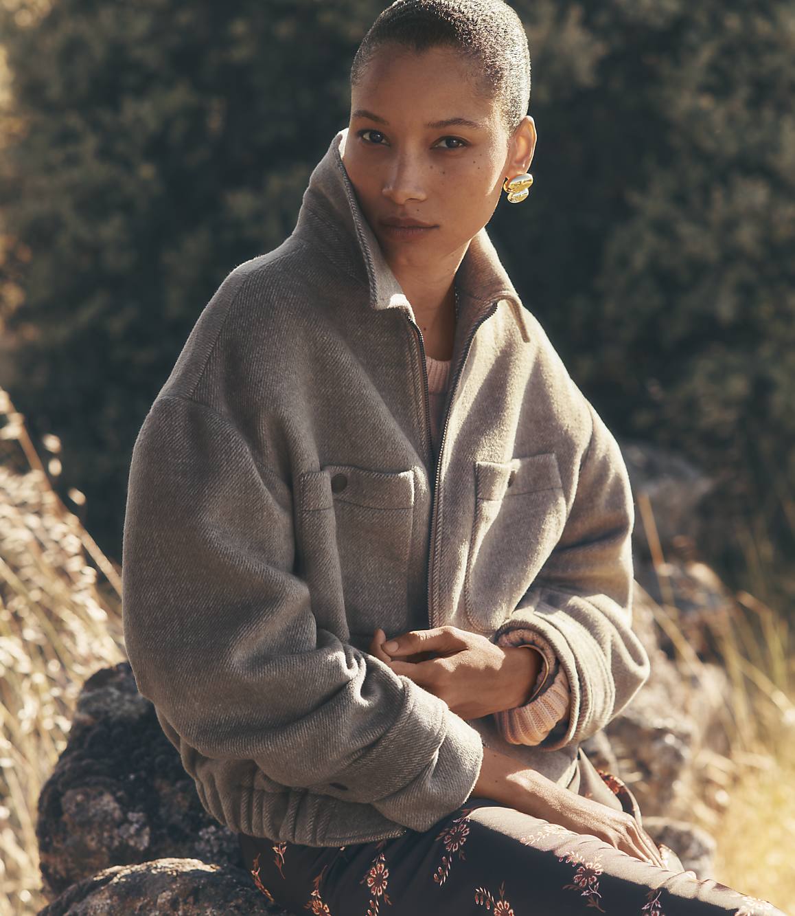A woman with short hair and a neutral expression sits outdoors in the foreground, wearing a grey jacket and patterned pants, with a blurred natural background of trees and rocks.