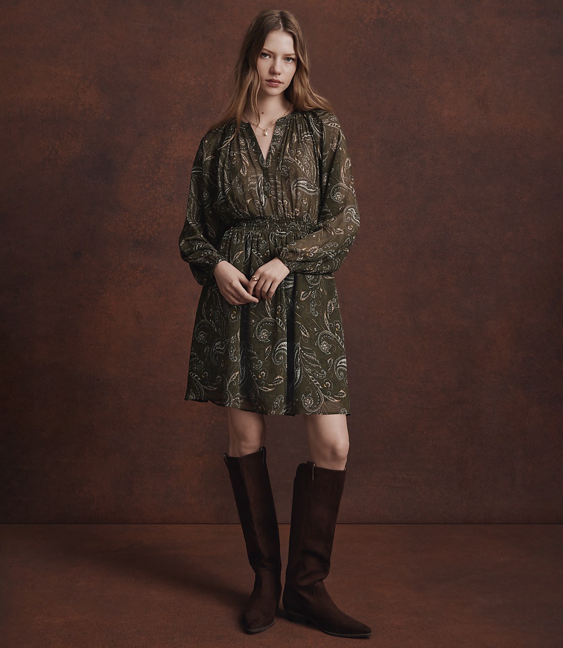 A woman in a patterned green dress and brown boots stands against a textured brown background.