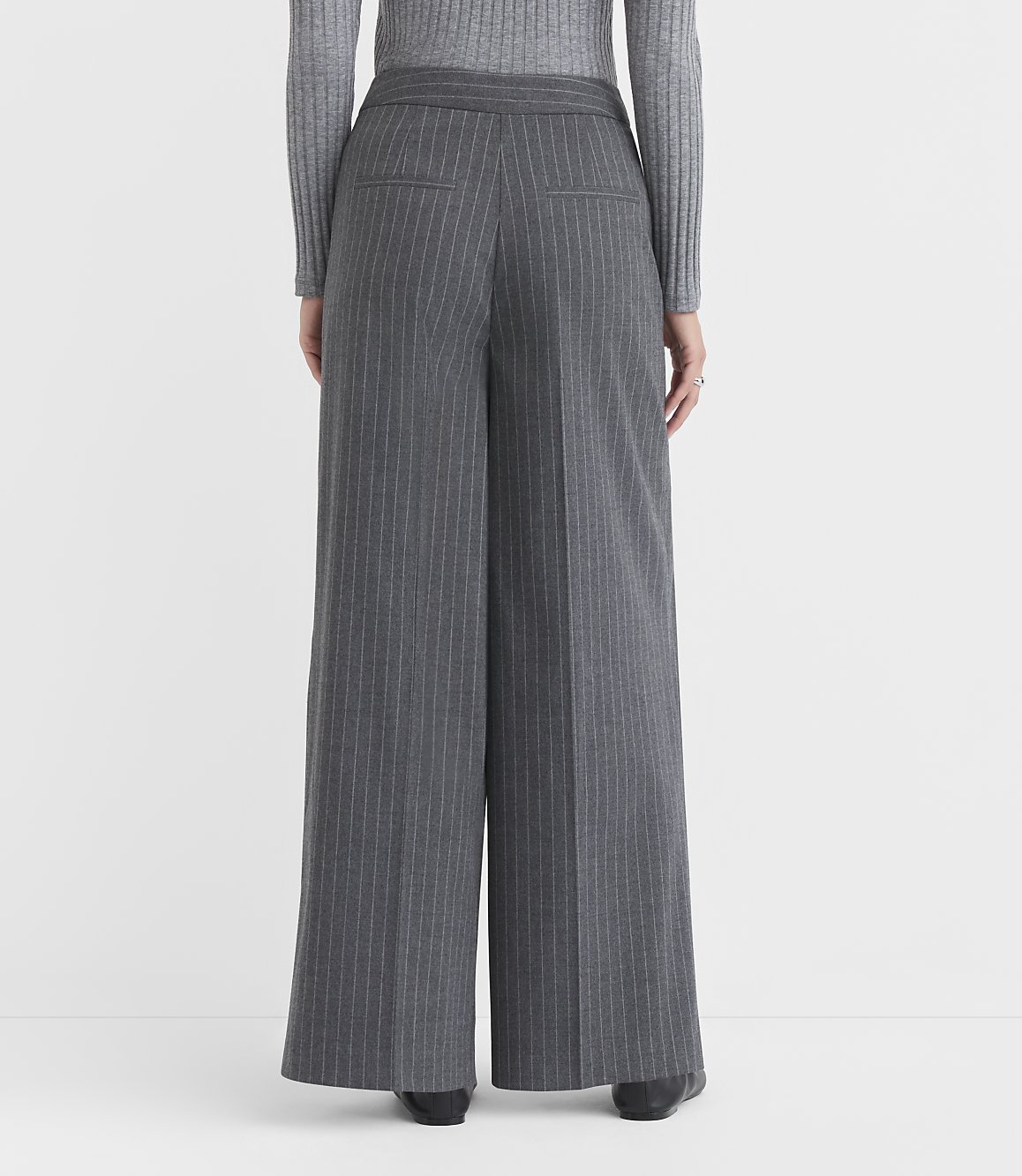 A person wearing gray pinstripe wide-leg pants and a gray sweater stands against a plain gray background.