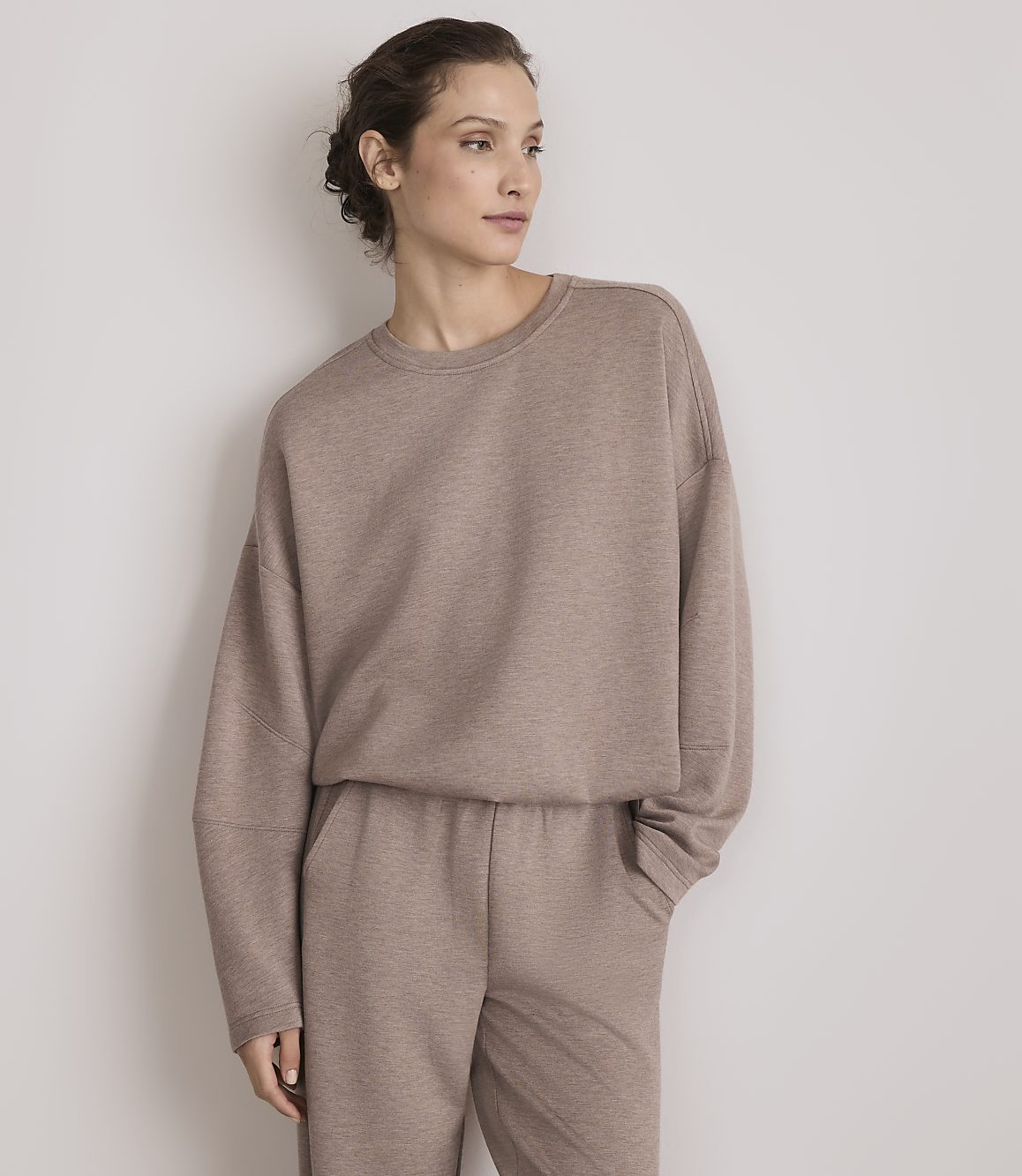 A woman in a taupe sweatshirt and matching pants stands against a plain, light gray background.