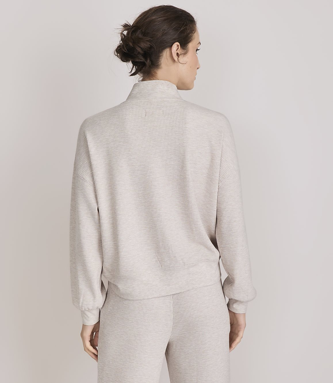 A woman in a beige mock-neck sweater and matching pants stands against a plain white background.