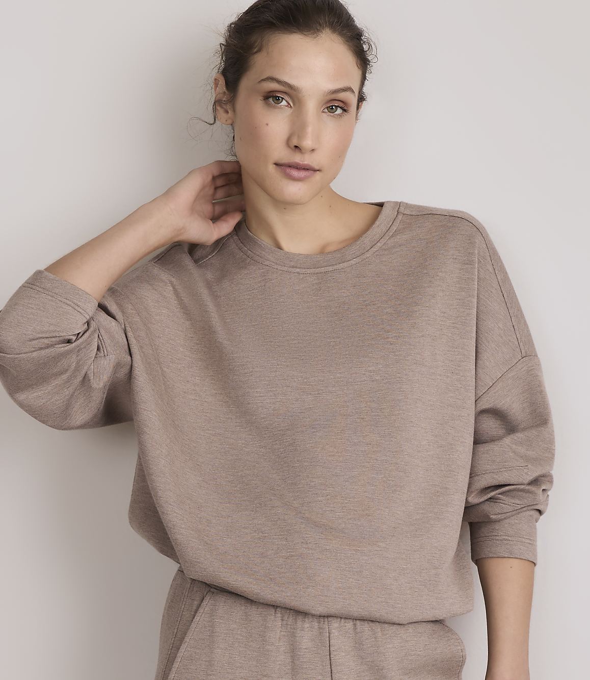 A woman in a taupe sweatshirt and matching pants poses against a plain, light-colored wall.