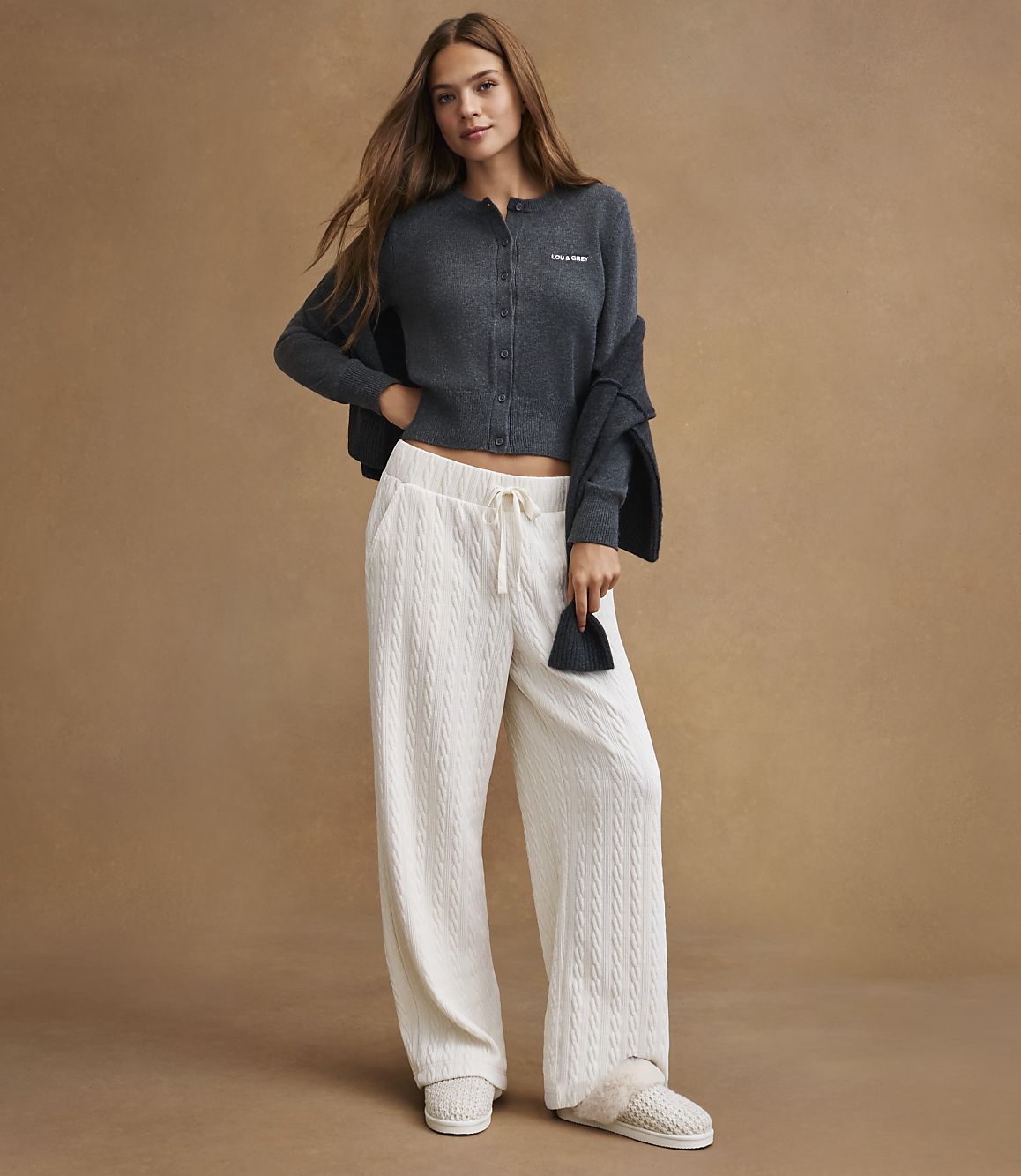 A young woman in a dark gray cardigan and white cable-knit pants stands against a plain brown background.