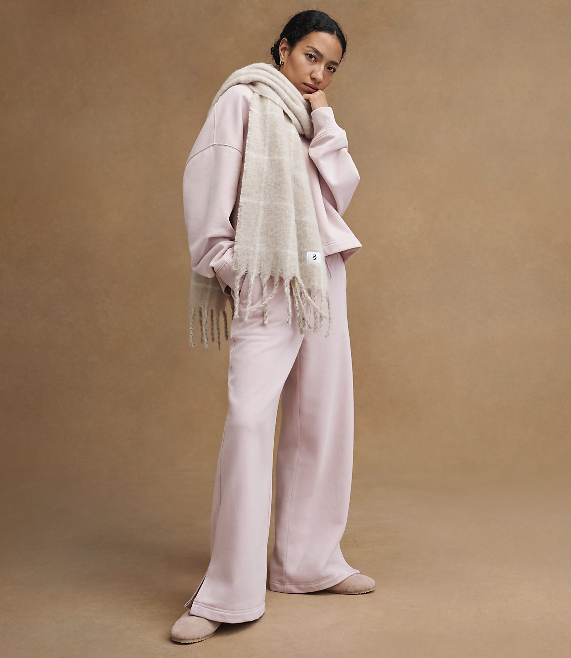 A woman in a pink loungewear set and a beige scarf stands against a plain brown background.