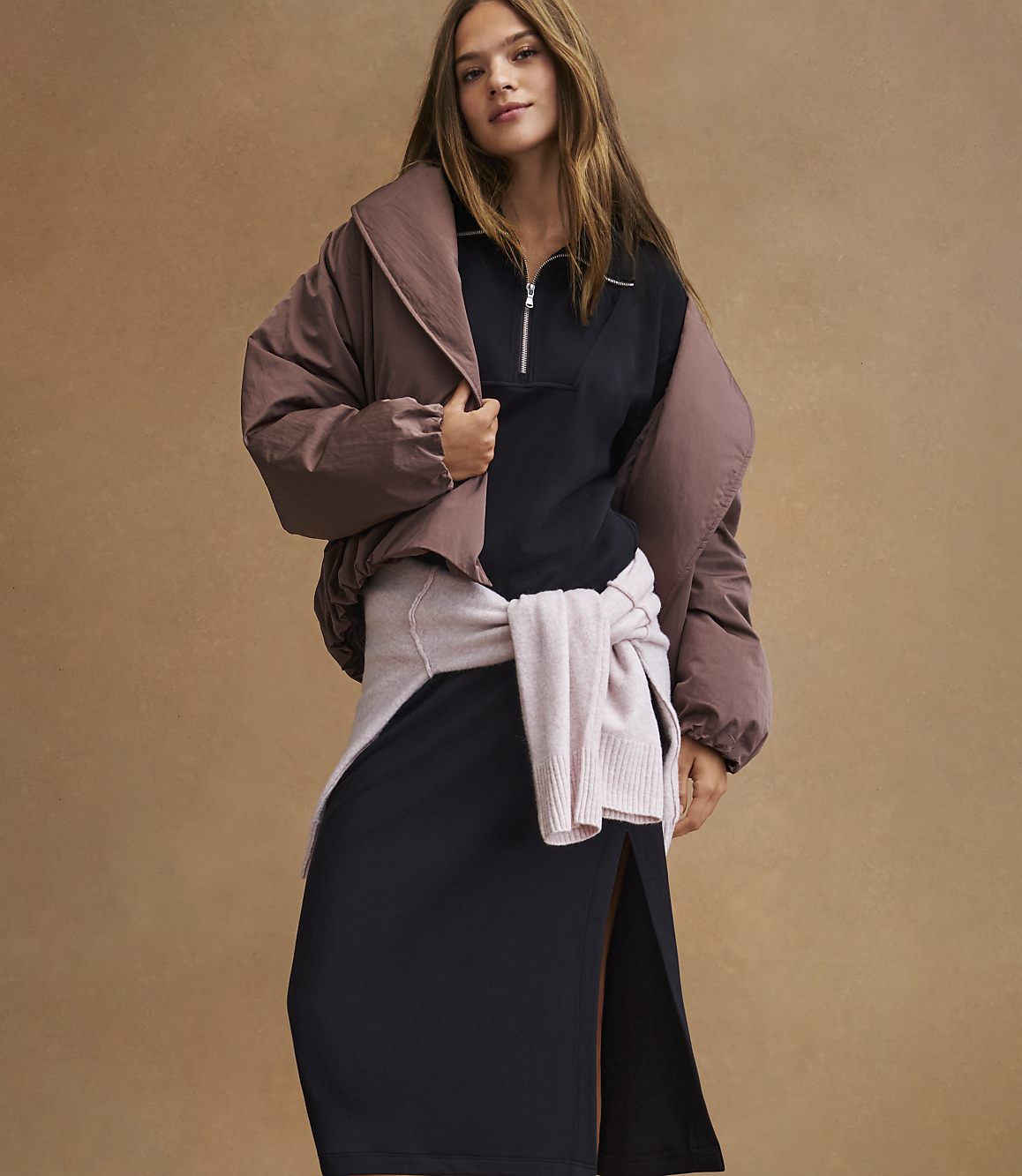 A woman in a black dress and a brown puffer jacket stands against a plain brown background.