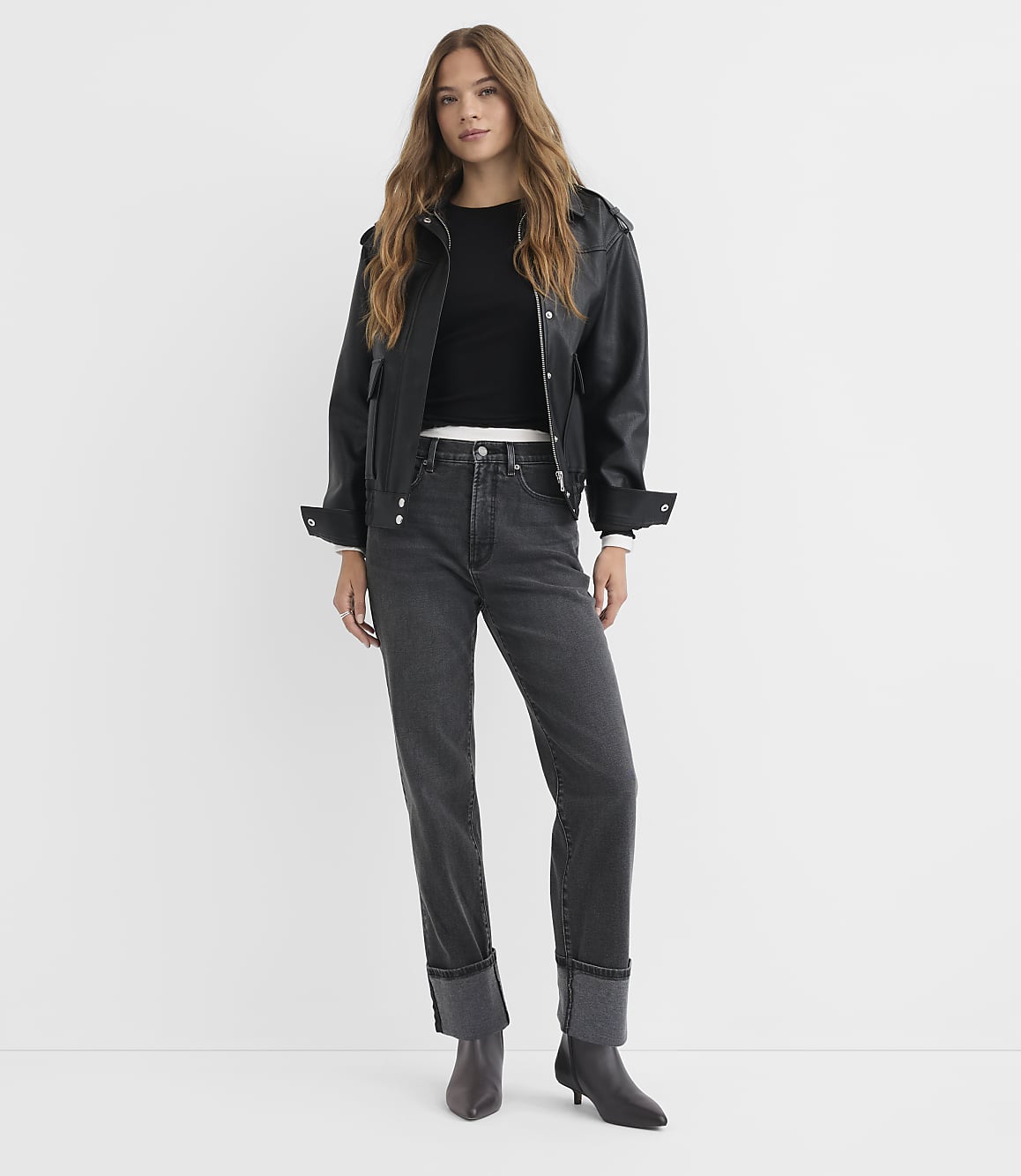 A woman in a black leather jacket and dark jeans stands against a plain white background.
