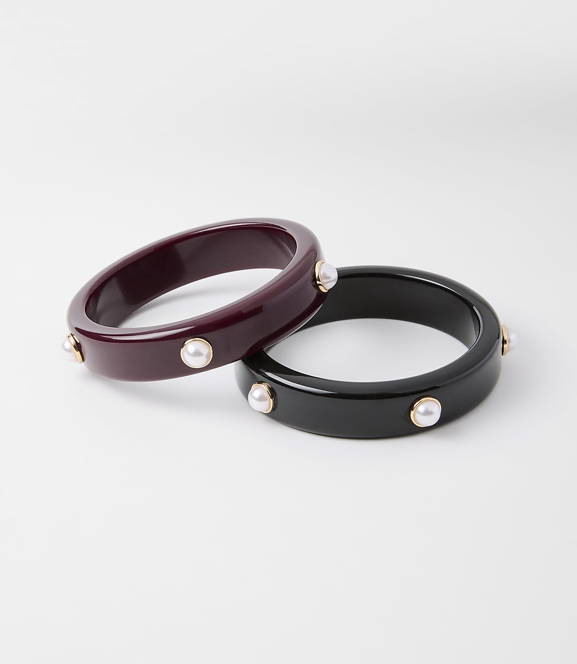 Two chunky bangles, one deep red and the other black, are adorned with small pearls and gold accents, set against a plain white background.