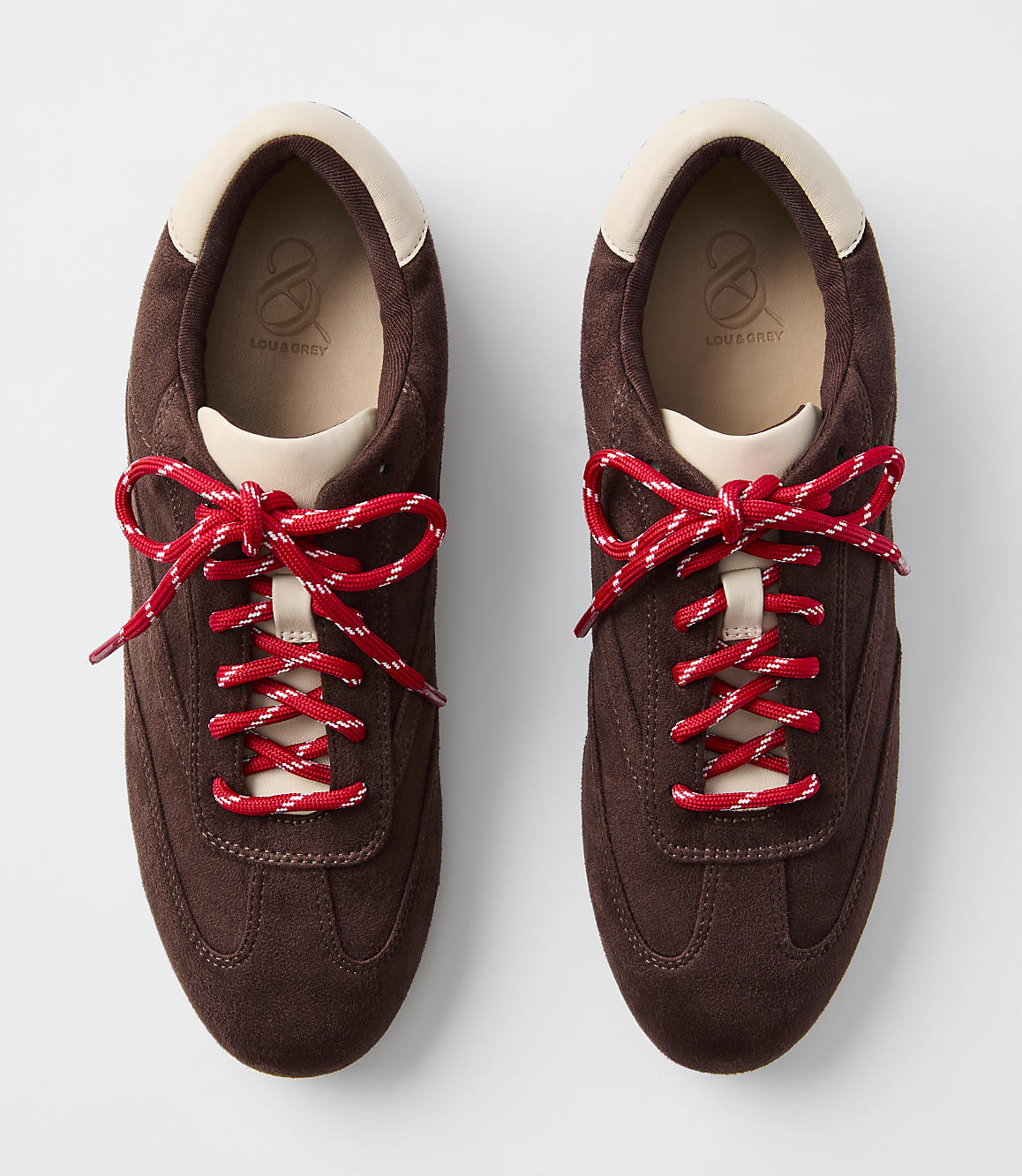 A pair of brown suede sneakers with red and white laces are displayed against a plain white background.