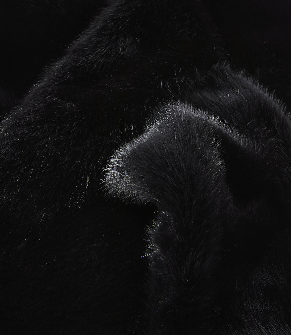 A close-up of black faux fur with soft folds in the foreground and a darker, less textured background.