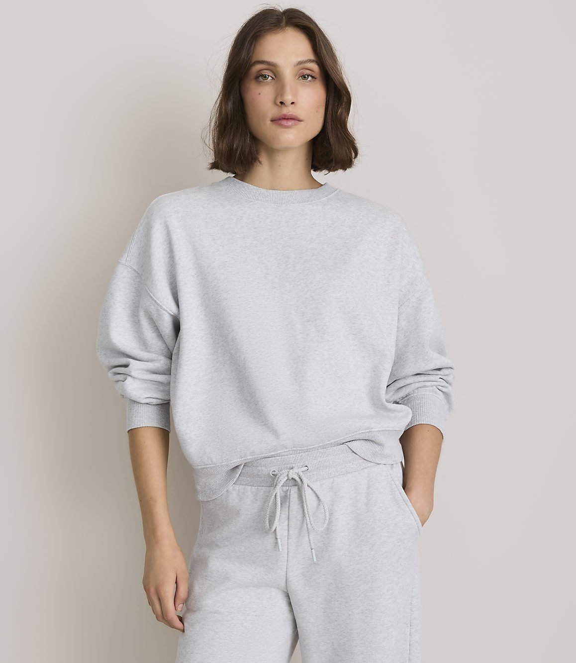 A woman in a grey sweatshirt and sweatpants stands against a plain, light grey background.