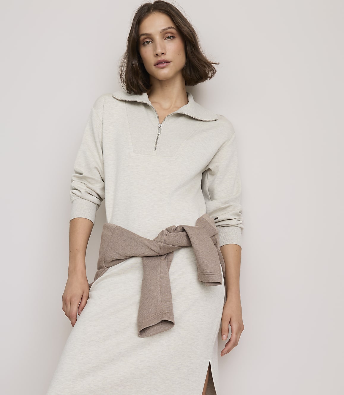 A woman in a light gray collared dress with a brown sweater tied around her waist stands against a plain, light-colored background.