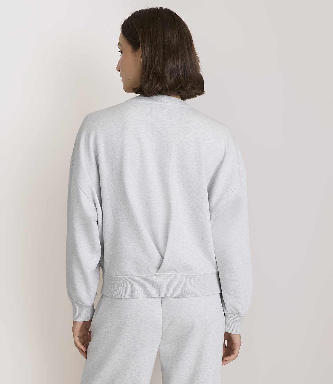 A woman in a grey sweatshirt and sweatpants stands against a plain white background.