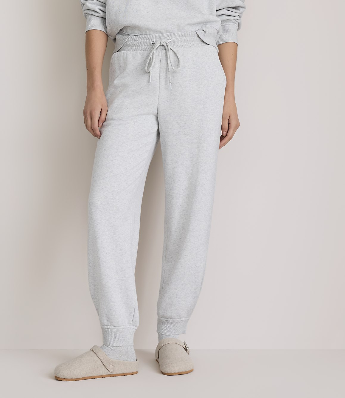 A person wearing light grey sweatpants and slippers stands against a plain, light grey background.
