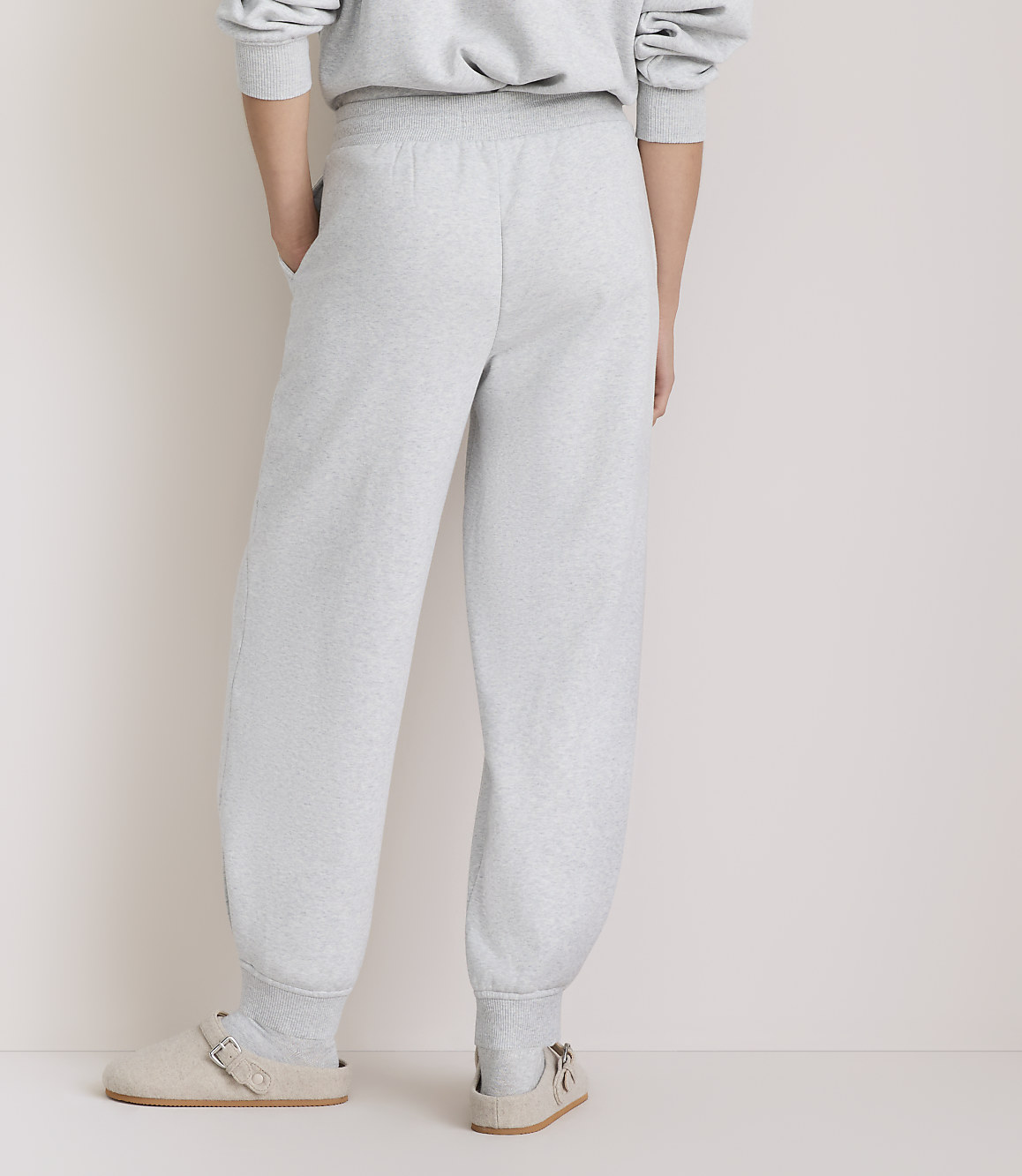 A person wearing light gray sweatpants and clogs stands against a plain, light-colored background.