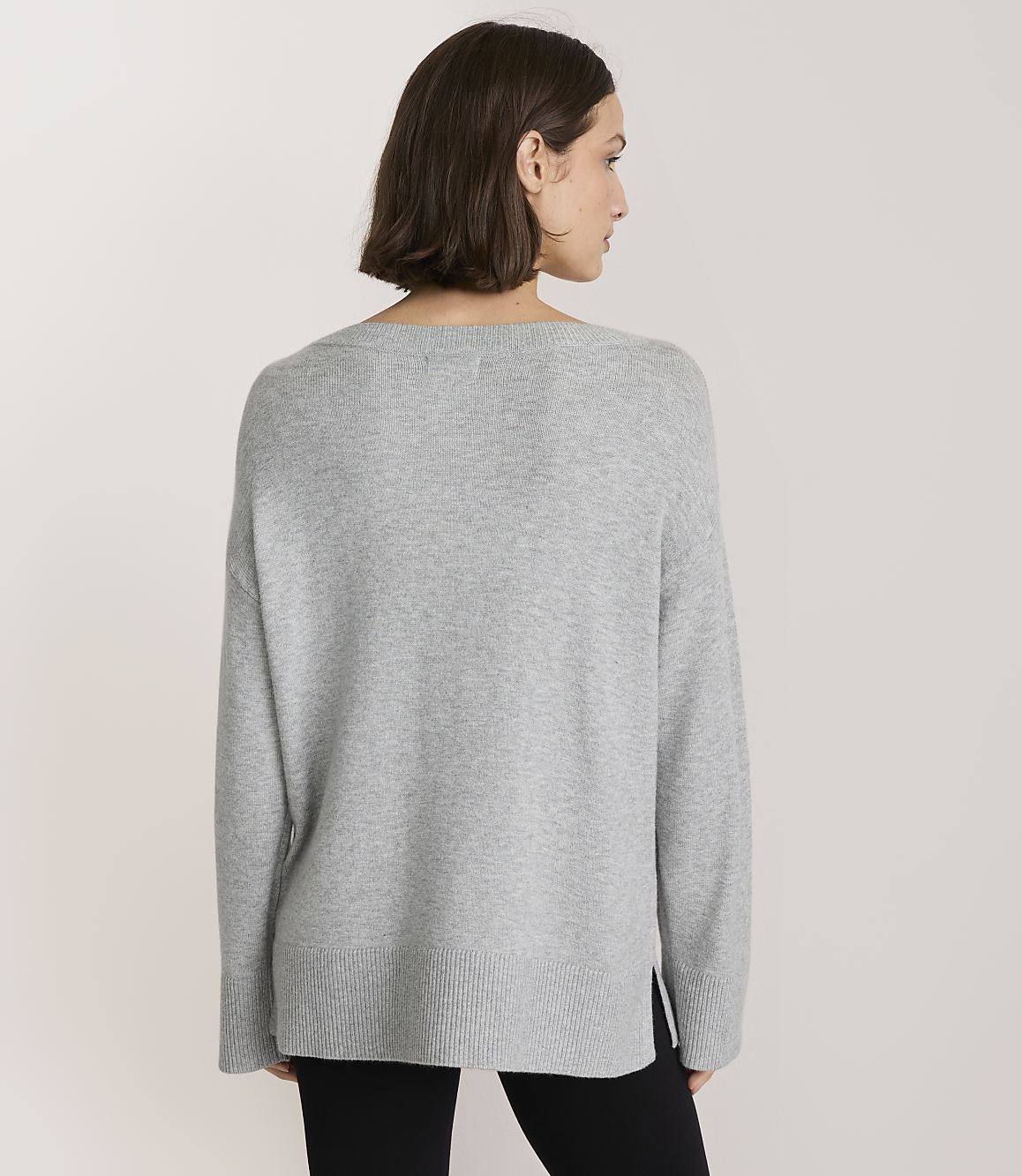 A woman in a grey sweater stands against a plain, light grey background.